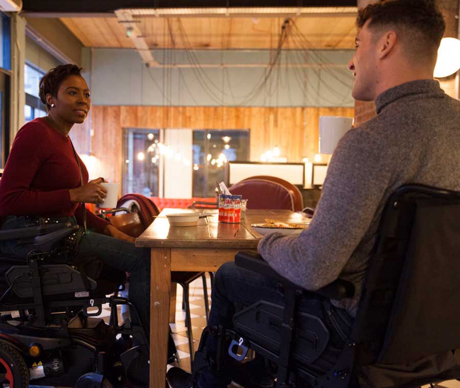 Q200R Powerchair. 2 people using their powerchair raised feature while sitting having a coffee at a table